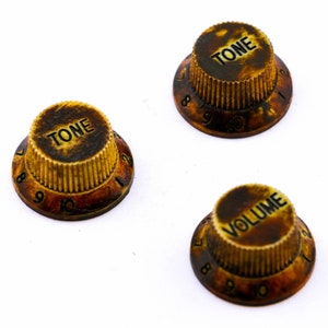 May include: Three vintage-style guitar knobs with a worn, aged look. The knobs are labeled "TONE" and "VOLUME" and have numbers 1-10 around the edges.