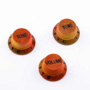 Könnte beinhalten: Drei orangefarbene und hellbraune Gitarren-Regler. Zwei Knöpfe sind mit "TONE" und einer mit "VOLUME" beschriftet. Jeder Knopf hat eine nummerierte Skala von 7 bis 10. Die Knöpfe sind vor weißem Hintergrund.