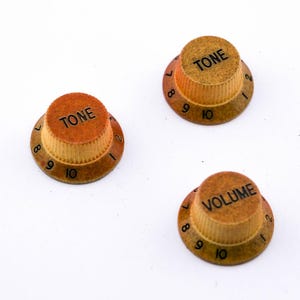 Könnte beinhalten: Drei Gitarrenknöpfe mit den Zahlen 1-10 und den Worten "TONE" und "VOLUME" darauf gedruckt. Die Knöpfe sind braun und gelb.
