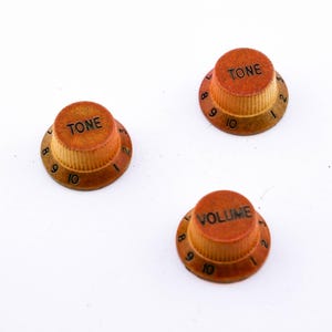Könnte beinhalten: Drei Gitarren-Regler im Vintage-Stil. Zwei Knöpfe sind mit "TONE" und einer mit "VOLUME" beschriftet. Die Knöpfe sind rotbraun und tragen die Zahlen 8, 9 und 10. Die Knöpfe befinden sich auf weißem Hintergrund.