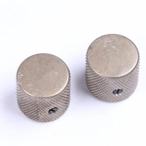 Op de afbeelding: Twee zilverkleurige, cilindrische knoppen met een getextureerde, diamantvormige grip. Elke knop heeft een platte bovenkant en een klein schroefgat. Deze knoppen zijn waarschijnlijk voor muziekinstrumenten of elektronische apparatuur.