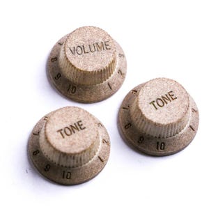 Könnte beinhalten: Drei beige Gitarrenknöpfe, zwei mit der Aufschrift "TONE" und einer mit "VOLUME". Jeder Knopf hat Zahlen von 1 bis 10 am Rand. Die Knöpfe bestehen aus einem strukturierten Material.