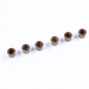 May include: A collection of six small, cylindrical metal bushings arranged in a row. Each bushing has a bronze-colored interior and a silver-toned exterior, set against a plain white background. These components appear to be for mechanical or engineering applications.
