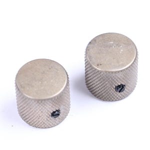 May include: Two silver-colored, cylindrical knobs with a textured, diamond-patterned surface. Each knob has a flat top and a small, black screw. The knobs are likely for a musical instrument or electronic device.