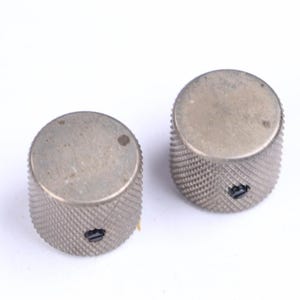 May include: Two silver-colored control knobs with a textured, ridged surface. The knobs have a cylindrical shape with a flat top and a small slot. The knobs are likely for an electronic device, such as a guitar amplifier or stereo receiver.