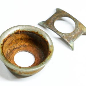 May include: Two antique metal parts with a weathered appearance. One is a bowl-shaped piece with a central hole and a rusty brown interior. The other is a flat, angular piece with a circular opening and pointed edges, showing signs of age.