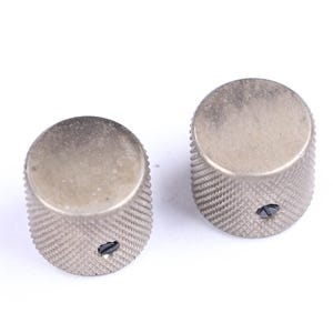 May include: Two silver-colored guitar knobs with a textured, ridged surface. Each knob has a small, dark screw visible on the side. The knobs are designed for use on electric guitars or similar instruments.