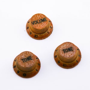Pode incluir: Três botões de guitarra com números e texto pretos. Os botões são marrons com uma superfície texturizada. O texto nos botões diz "VOLUME" e "TONE".