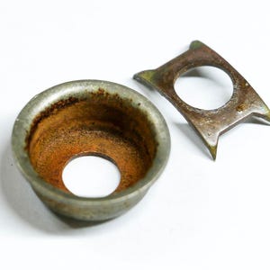 May include: Close-up of two antique metal objects with a weathered appearance. One is a circular, cup-shaped piece with a central hole and rust-colored interior. The other is a flat, angular piece with a central hole and pointed ends.