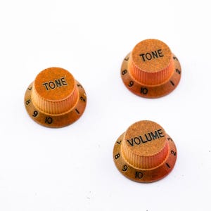 Könnte beinhalten: Drei Gitarrenknöpfe mit einem braunen und orangen Farbverlauf. Die Knöpfe sind mit "TONE" und "VOLUME" sowie den Zahlen 1 bis 10 beschriftet.