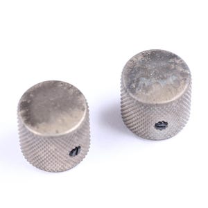 May include: Two silver-toned, cylindrical knobs with a textured, ridged surface. Each knob has a small, dark hole on the side. The knobs are likely for a musical instrument or electronic device.