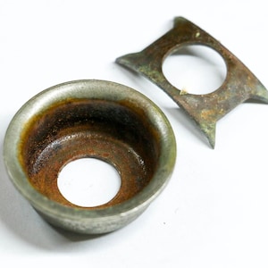 May include: Two antique metal objects with a weathered appearance. One is a small, round, cup-shaped piece with a central hole and a rusty interior. The other is a flat, angular piece with a circular opening and pointed edges.