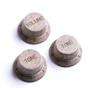 Könnte beinhalten: Drei strukturierte, hellbraune Gitarrenknöpfe mit den Aufschriften "VOLUME" und "TONE". Jeder Knopf hat eine kreisförmige Basis mit Zahlen von 0 bis 10. Die Knöpfe dienen zur Einstellung der Lautstärke und des Klangs einer E-Gitarre.