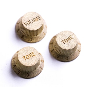 Könnte beinhalten: Drei beige Gitarrenknöpfe mit den Wörtern "VOLUME" und "TONE" aufgedruckt. Die Knöpfe haben Zahlen von 8 bis 10. Die Knöpfe bestehen aus einem strukturierten Material und sind vor einem weißen Hintergrund platziert.