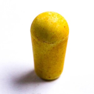 May include: A yellow, cylindrical object with a rounded top. The surface has a textured appearance with small, darker spots. The object is set against a white background, casting a soft shadow.