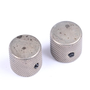 May include: Two silver-colored, cylindrical knobs with a textured, diamond-patterned surface. Each knob has a small, black screw visible on the side. The knobs show signs of wear and tear, with dark spots on the top.