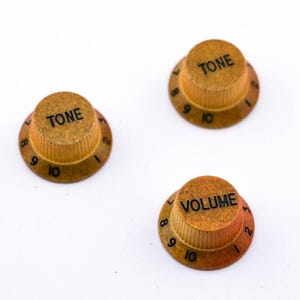 Könnte beinhalten: Drei Gitarrenknöpfe mit goldener Oberfläche und schwarzer Beschriftung. Die Knöpfe sind mit "TONE" und "VOLUME" beschriftet und haben die Zahlen 1 bis 10 am Rand.