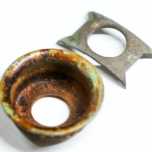 May include: An overhead shot shows two antique metal objects. One is a small, round, cup-shaped piece with a central hole, featuring brown, green, and orange hues. The other is a flat, angular piece with a circular opening and a weathered, gray-green finish.