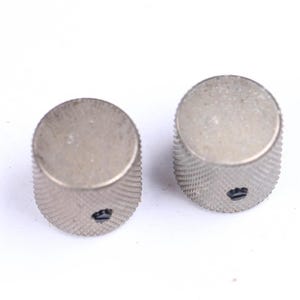May include: Two silver-colored, cylindrical knobs with a textured, ridged surface. Each knob has a small, dark emblem at the base. The knobs are likely for a musical instrument or electronic device.