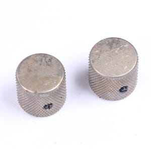 May include: Two silver-colored control knobs with a textured, ridged surface. The knobs have a cylindrical shape with a flat top and a small screw on the side. The knobs are likely for an electronic device.