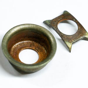 May include: Two antique metal objects. One is a small, round, cup-shaped piece with a central hole, showing a mix of green and brown tones. The other is a flat, angular piece with a central hole and pointed edges, also with a weathered appearance.