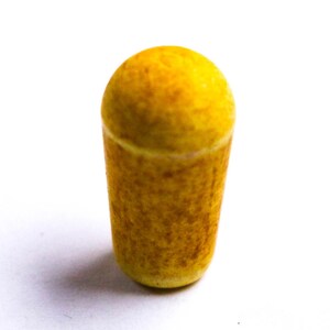 Könnte beinhalten: Ein zylindrischer Flaschenverschluss mit abgerundeter Oberseite. Der Verschluss ist überwiegend gelb mit einem strukturierten, gesprenkelten Aussehen. Die Basis ist ein hellerer Gelbton.