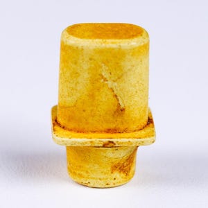 May include: A cylindrical bottle stopper with a square base and a rounded top. The stopper is a warm yellow color with darker brown markings, suggesting an aged or rustic appearance. The design is simple and functional.