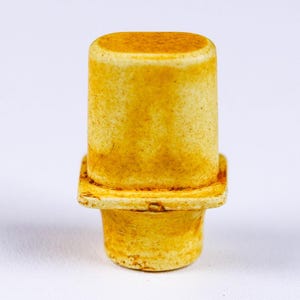 May include: A close-up of a yellow bottle stopper. The stopper has a cylindrical top, a square middle section, and a flared base. The color is a warm, aged yellow, with subtle variations in tone. The object is set against a plain white background.