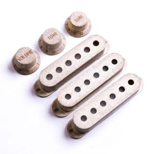Könnte beinhalten: Ein Satz Gitarrenteile, darunter drei Tonabnehmerabdeckungen und drei Regler. Die Knöpfe sind mit "VOLUME" und "TONE" beschriftet und mit Zahlen versehen. Die Teile haben eine strukturierte, hellbraune Oberfläche.