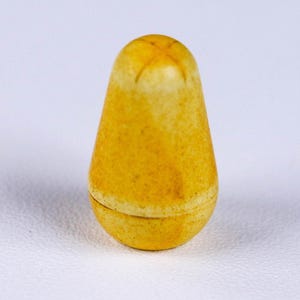 May include: A small, yellow, pear-shaped object with a cross-shaped design on the top. The object has a two-part construction, with a base and a top section. The surface has a slightly textured appearance.