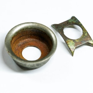 May include: Close-up of two antique metal components. One is a circular, cup-shaped piece with a rusty interior. The other is a flat, angular piece with a central hole and a weathered, green-tinged surface. Both show signs of age.