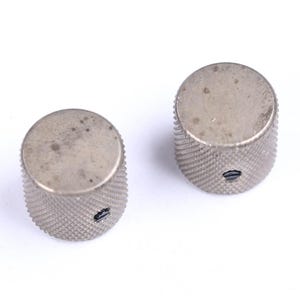 May include: Two silver-colored, cylindrical knobs with a textured, diamond-patterned grip. Each knob has a flat top and a small, oval-shaped hole. The knobs appear to be made of metal and are likely used for electronic equipment.