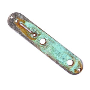 May include: A distressed metal guitar component, rectangular with rounded ends. The metal shows a mottled pattern of green, brown, and grey, and has several holes. The part has an aged or weathered appearance.