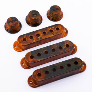 Könnte beinhalten: Eine Sammlung von Gitarrenteilen, darunter drei Klangregler, ein Lautstärkeregler und drei Tonabnehmerabdeckungen. Die Knöpfe und Abdeckungen haben ein Schildpattmuster mit braunen und schwarzen Farbtönen. Der Lautstärkeregler ist mit "VOLUME" beschriftet, die Klangregler mit "TONE".