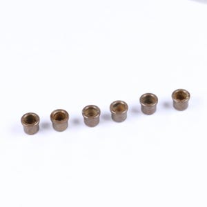 May include: A collection of seven small, cylindrical bronze bushings arranged in a row. Each bushing has a hollow center and a slightly flared top, with a smooth, metallic finish. The bushings are set against a plain white background.