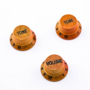 Könnte beinhalten: Drei Gitarrenknöpfe mit schwarzen Zahlen und Beschriftung. Die Knöpfe sind orange und braun mit einer strukturierten Oberfläche. Die Knöpfe sind mit "TONE" und "VOLUME" beschriftet.