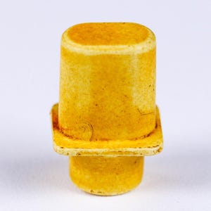May include: A close-up of a yellow bottle stopper with a cylindrical top and a square base. The stopper has a rounded bottom and a slightly textured surface. The color is a warm, aged yellow.