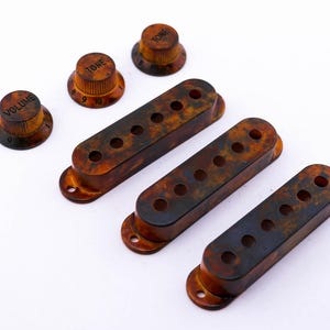 Può includere: Tre coperture per pickup di chitarra marroni e nere e tre manopole per una chitarra. Le manopole sono etichettate "Volume" e "Tono".