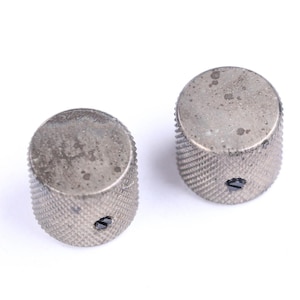 May include: Two silver-colored, cylindrical knobs with textured sides and a small hole. The knobs have a slightly worn appearance, suggesting use. These are likely replacement parts for musical instruments or electronics.