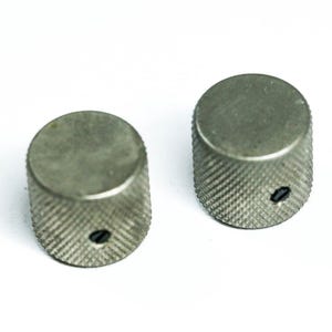 Könnte beinhalten: Zwei silberfarbene, zylindrische Knöpfe mit geriffelter, rautenförmiger Grifffläche. Jeder Knopf hat eine flache Oberseite und ein kleines, dunkles Loch. Diese Knöpfe sind wahrscheinlich für den Einsatz an elektronischen Geräten bestimmt.