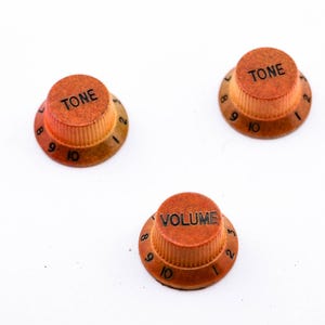 Könnte beinhalten: Drei orangefarbene Gitarrenknöpfe mit schwarzer Beschriftung. Die Knöpfe sind mit "TONE" und "VOLUME" beschriftet.