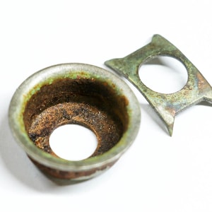 May include: Two antique metal components with a weathered appearance. One is a circular piece with a central hole and a green-tinged rim. The other is a flat, angular piece with a circular opening and pointed ends. Both show signs of age.