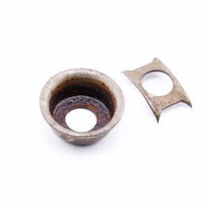 May include: Two antique metal components, likely from a larger mechanism, rest against a white background. One is a small, circular cup with a central hole and dark interior. The other is a small, irregular metal piece with a circular opening.