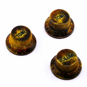 Könnte beinhalten: Drei vintage goldfarbene Gitarrenknöpfe mit schwarzer Beschriftung. Die Knöpfe sind mit "TONE" und "VOLUME" beschriftet und haben die Zahlen 7 bis 10, die um die Ränder gedruckt sind.