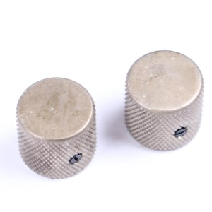 May include: Two silver-colored, cylindrical knobs with a textured, ridged surface. Each knob has a flat, circular top and a small screw visible on the side. The knobs are likely for a musical instrument or electronic device.