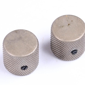 May include: Two silver-toned control knobs with a textured, ridged surface. Each knob has a flat top and a small, black screw. The knobs are likely for an electronic device, such as a guitar or amplifier.