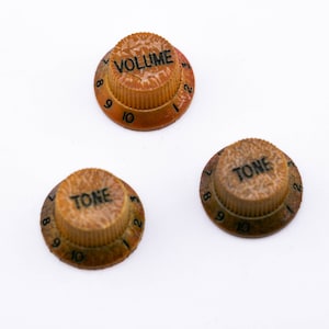Könnte beinhalten: Drei Vintage-Gitarrenknöpfe mit den Zahlen 1-10, die um den Rand gedruckt sind. Die Knöpfe sind braun und haben die Wörter "VOLUME" und "TONE" aufgedruckt.