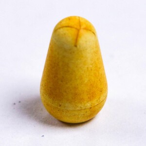 May include: A yellow, pear-shaped object with a cross-shaped design on top. The object has a textured surface and a rounded base. The color is a mix of yellow and brown, with darker speckles throughout.