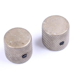 May include: Two silver-colored, cylindrical knobs with a textured, diamond-patterned surface. Each knob has a small, dark screw visible on the side. The knobs are likely for a musical instrument or electronic device.