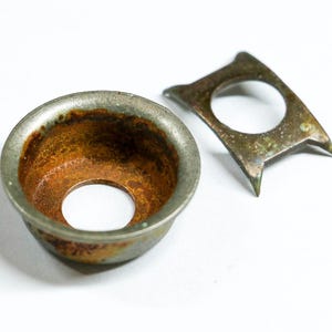 May include: A close-up of two aged metal objects. One is a small, round, bowl-shaped piece with a central hole and a rusty interior. The other is a flat, angular piece with a circular opening and pointed edges. Both show signs of age.